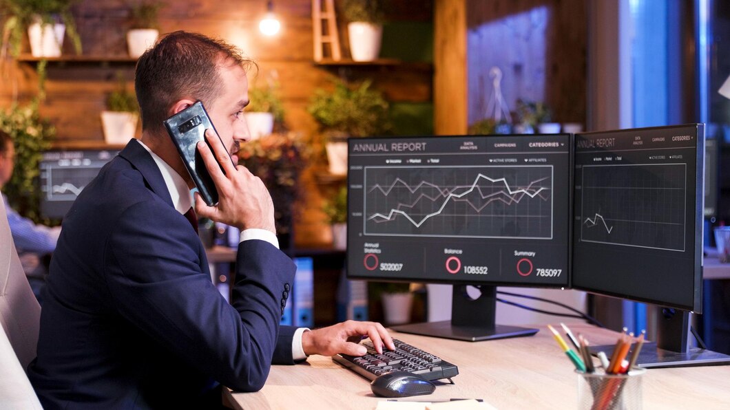 Homme en costume au téléphone, travaillant sur des graphiques financiers sur deux écrans.